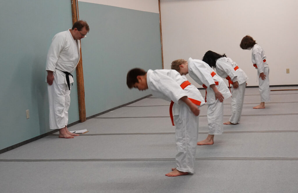 three children in Aikido dogis and orange belts are standing in a line bowing to their sensei and